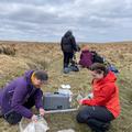 two people in the foreground of a peatland stream, sitting next to river sampling equipment, with a group of colleagues in the background