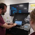 Photograph of a member of staff, wearing a red tshirt, showing student a display on a screen. The screen is connected to a mass spectrometry instrument housed inside a suitcase.