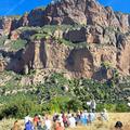 Students in bright coloured clothes sat in a group in a sunny field with a large rock face behind them.