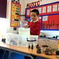 A photograph of a young child pouring water into a large bucket as part of a science experiment. They are standing in a school classroom.