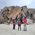 Photograph of demonstrators stood in front of a large rock on a beach. They are facing away from the camera and wearing outdoor gear.