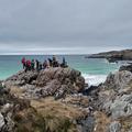 Photograph from a distance of a large group of students stood on a huge rock overlooking a blue-green sea and white beach