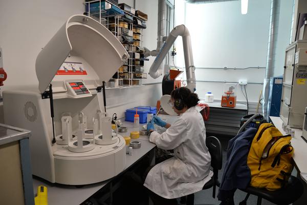 Student working using a brush on a sample at a lab bench next to an open ball mill. She is wearing gloves, a lab coat, and ear defenders. Ventilation ducts and storage shelves are visible in the background.