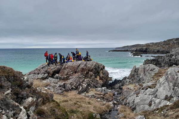 Students on the Assynt Field Class 1 Photograph from a distance of a large group of students stood on a huge rock overlooking a blue-green sea and white beach