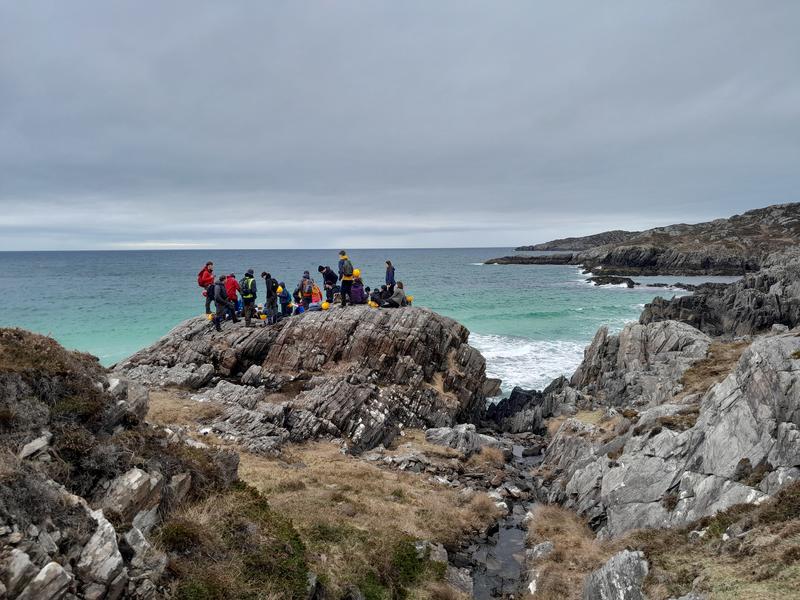 Students on the Assynt Field Class 1 Photograph from a distance of a large group of students stood on a huge rock overlooking a blue-green sea and white beach