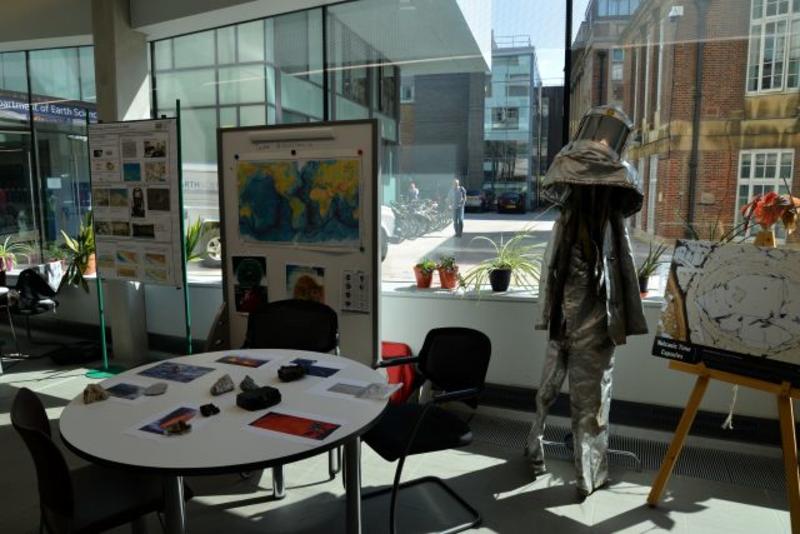 A display of volcanology equipment at an Open Day. The equipment includes a volcanologist suit and a table of samples. The display is situated in front of some large glass windows.