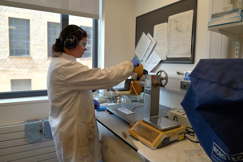 Lab manager Emily feeds a sample into a Frantz isodynamic magnetic separator. She is wearing a lab coat, gloves and hearing protection.