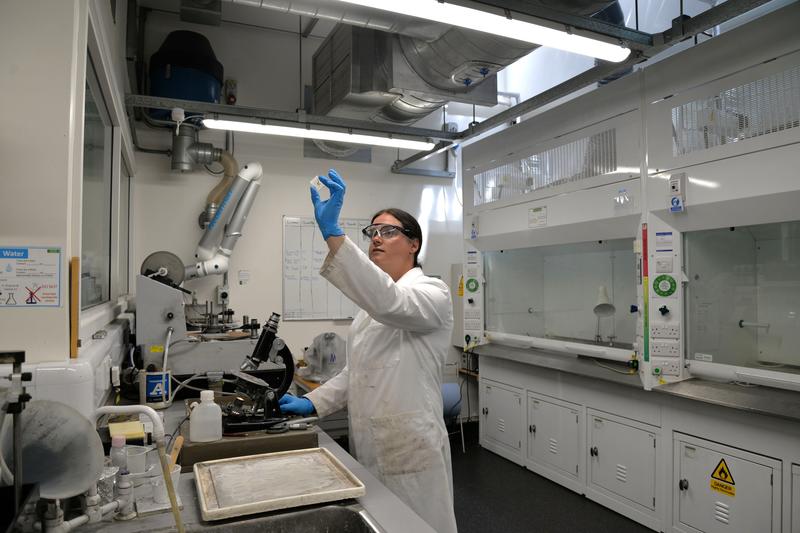 Lab manager Emily holds a thin section up to the light in the thin sections lab. She is wearing a lab coat and gloves. Other equipment including fume cupboards is visible in the background.