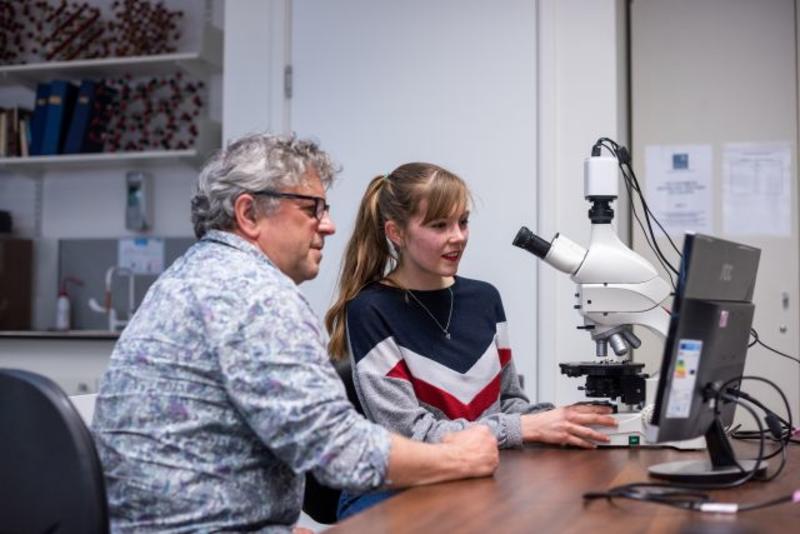 Photograph of a student and supervisor sat next to each other working on a large white microscope. There is a computer screen in front of them. 