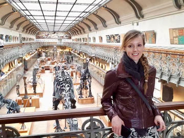Photograph of Erin Saupe standing on a balcony in a museum with exhibits behind her