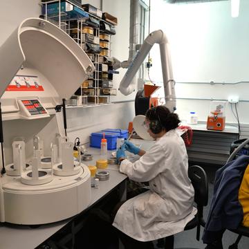 A student in a lab coat and hearing protection works at a bench in the crushing lab. In the background there is a large crushing machine, with sieves and sample containers also visible.