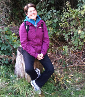 Photograph of Elizabeth Crowley. She is wearing a purple jacket and black trousers, has short hair, and is sat on a tree stump smiling at the camera.