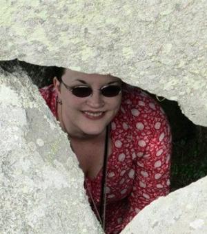 Photograph of Emma Brown. She is wearing a red spotty dress and sunglasses and peering through a gap in some rocks.