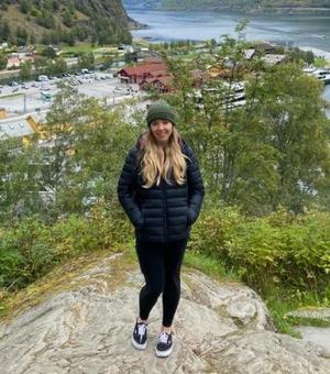 Photograph of Louisa Bailey. She is standing on a large rock with a view of a village in a valley behind her. She is wearing a black outfit with a green beanie.